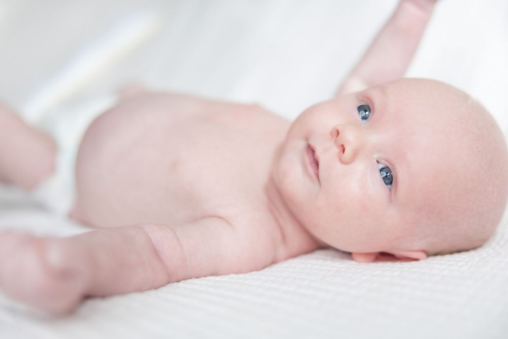 A baby with bright blue eyes lies on white bedding looking up at the camera.