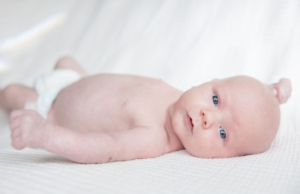 Newborn baby with soft rosy skin lies on white blanket while looking to the side.