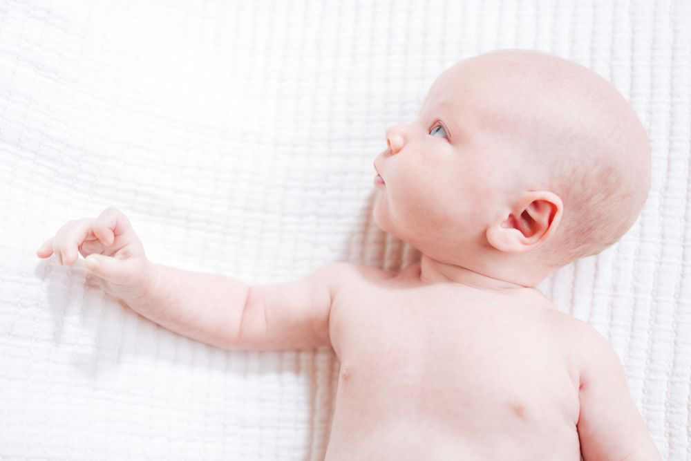 Newborn baby lying on white bedding with arm outstretched against soft textured background.