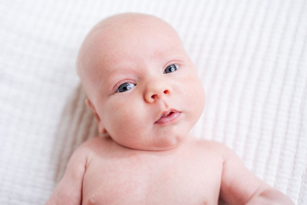 A baby with bright eyes and smooth skin lying on white textured fabric looking at the camera.