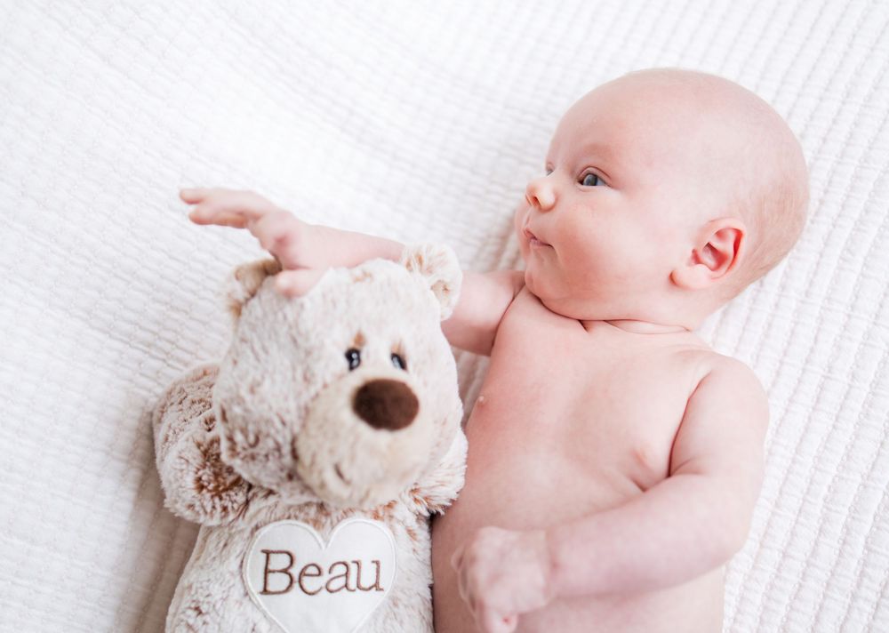 A baby lying on white bedding reaches out to touch a plush teddy bear with 'Beau' embroidered on it.