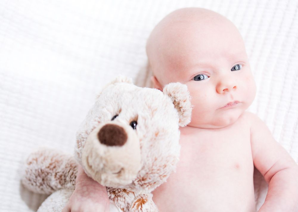 Sweet baby lies next to plush teddy bear on white blanket with dreamy expression.