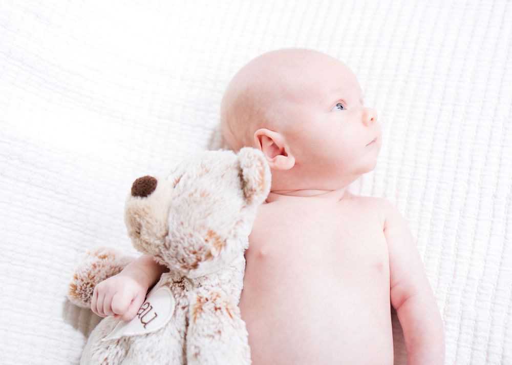A baby lying on white bedding next to a plush teddy bear toy.