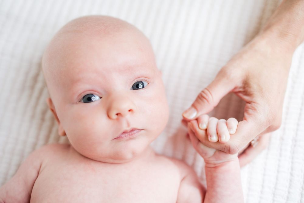 A baby lying on white blanket holding an adult's finger while looking at camera with wide eyes.