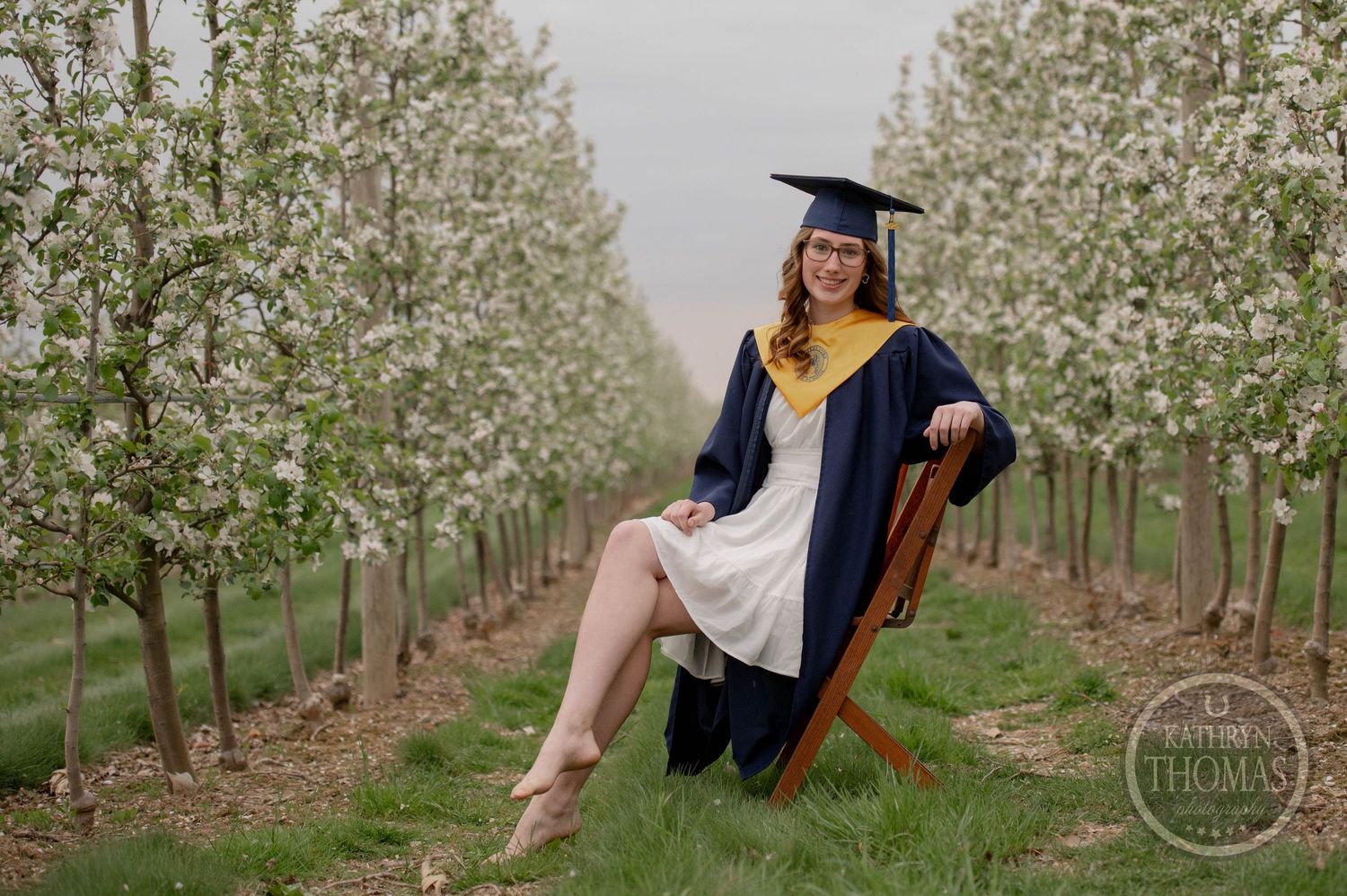 Graduate in cap and gown poses in wooden chair among blooming orchard trees on spring day.