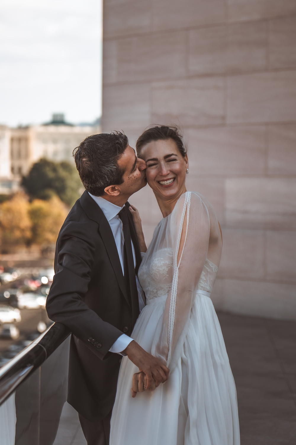 A romantic couple in formal wedding attire share an intimate moment on a balcony during golden hour.