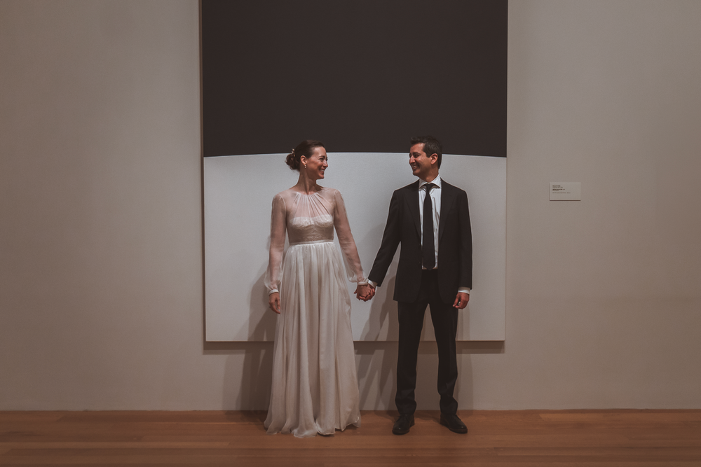 A couple on their wedding day holds hands while standing in front of a minimalist white wall in an indoor space.