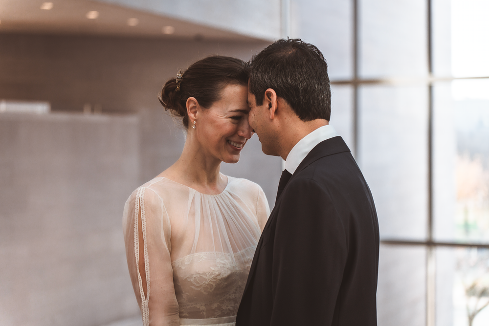 A romantic couple in wedding attire share an intimate moment by a window, with soft natural lighting surrounding them.