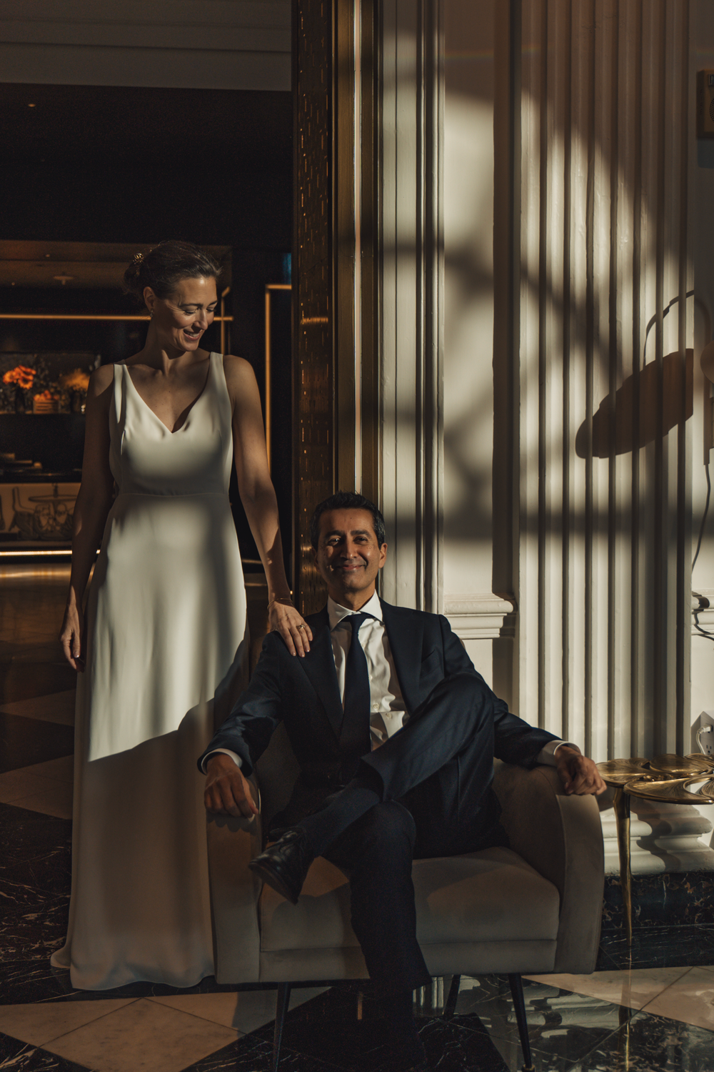 A couple in formal wedding attire poses elegantly in a luxurious indoor setting with dramatic window lighting.