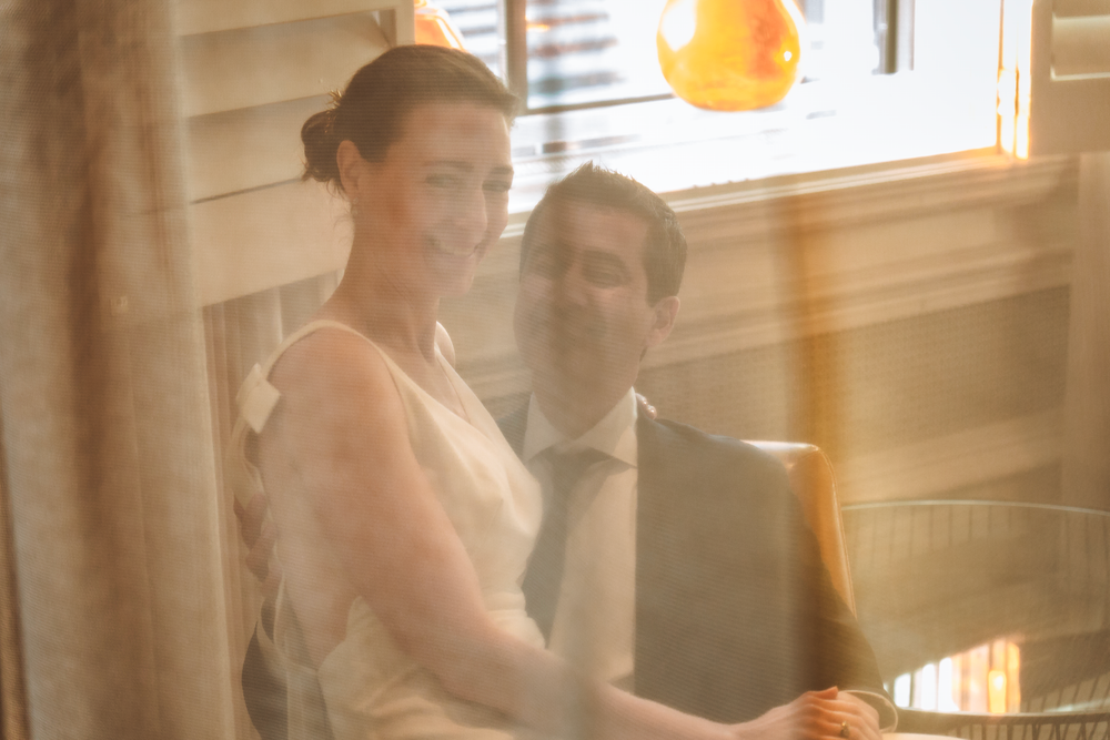 A bride and groom share an intimate moment behind a sheer curtain with soft natural light filtering through.