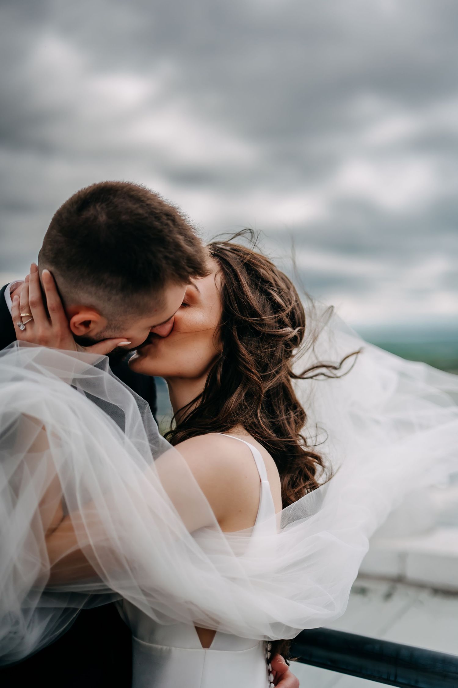 A tender intimate moment between newlyweds sharing a kiss outdoors with dramatic cloudy skies and flowing veil in the wind Grand Rapids