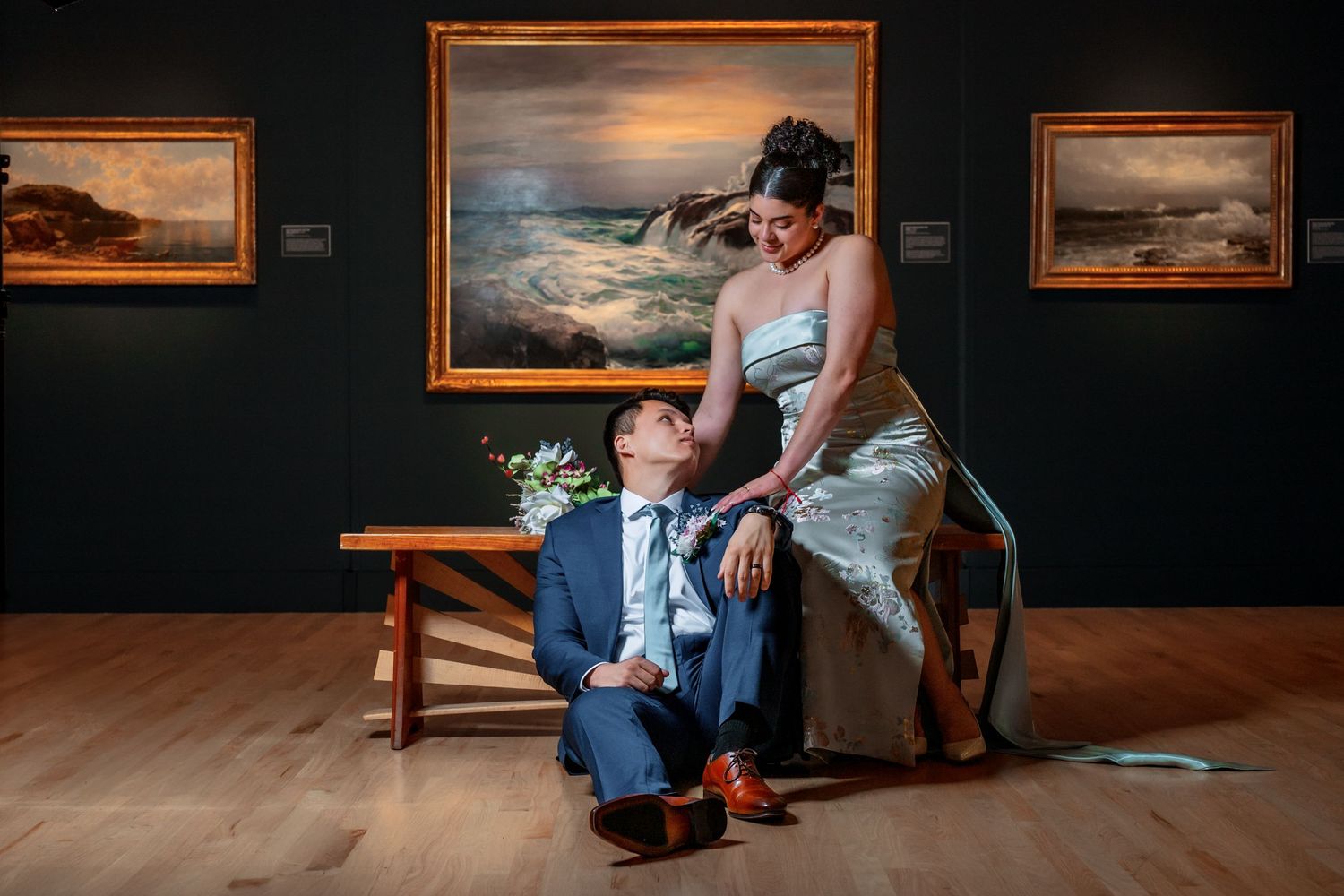 Couple sits on bench in art gallery with seascape paintings on dark walls.