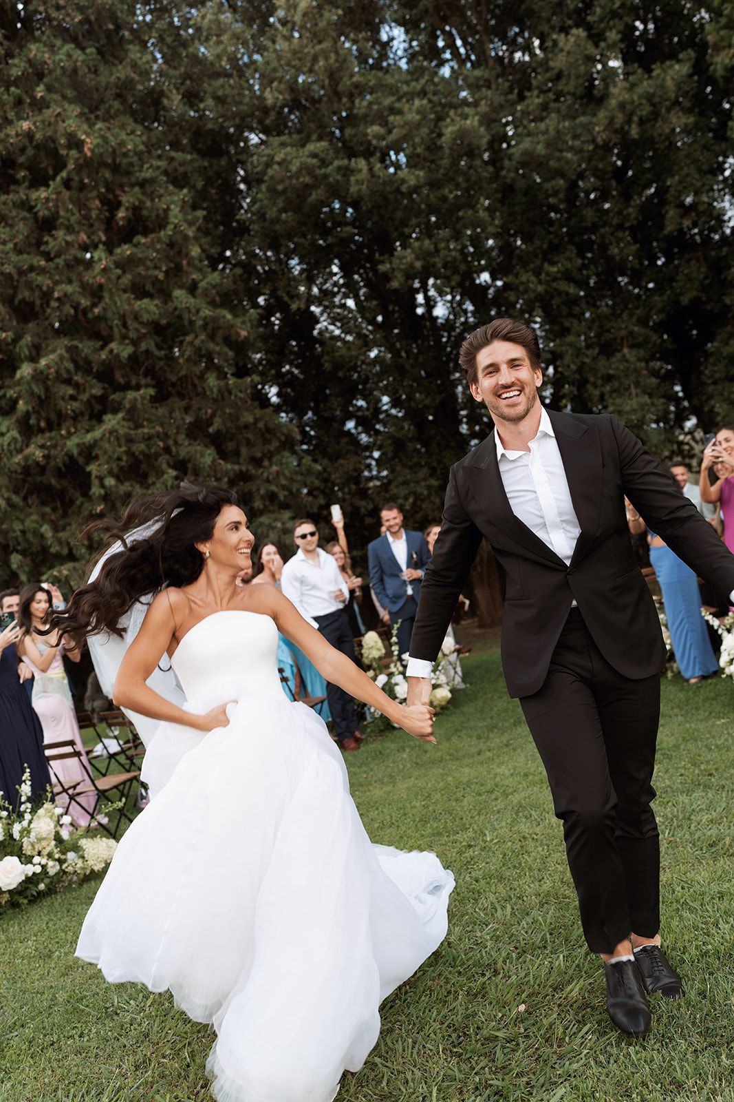 A bride in a white gown and groom in dark suit run joyfully across a grassy lawn at their outdoor wedding ceremony.