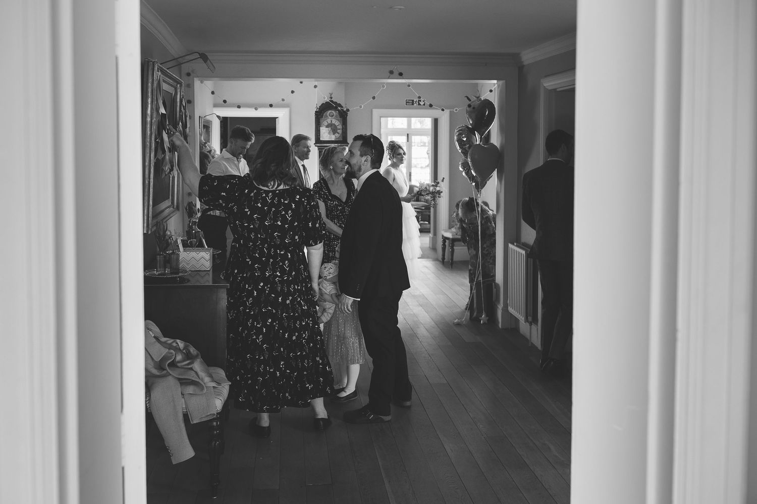 A vintage black and white photograph shows people gathered in a wood-floored hallway conversing and interacting.