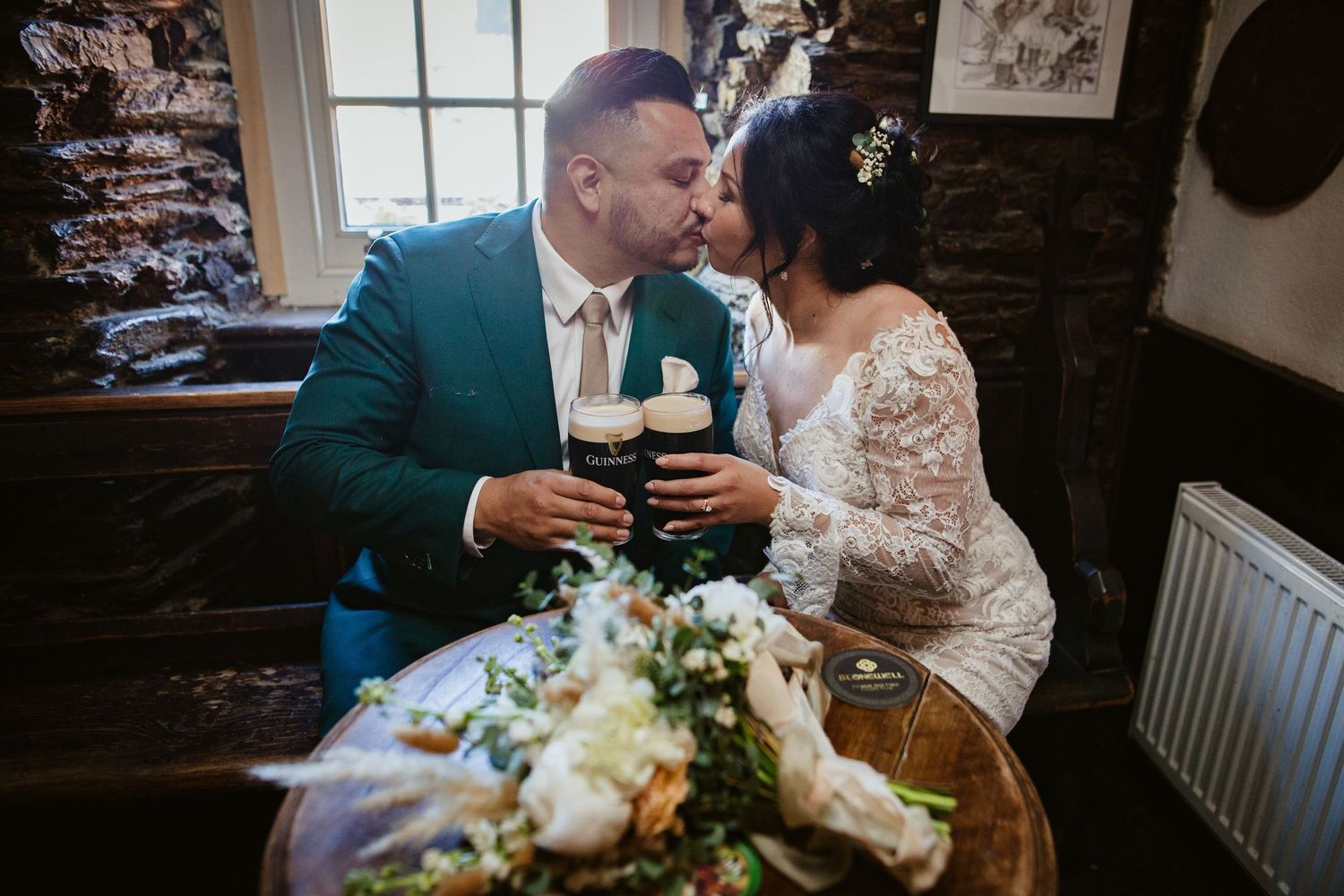 A couple in formal attire shares an intimate moment at a rustic pub table with wedding flowers and drinks.