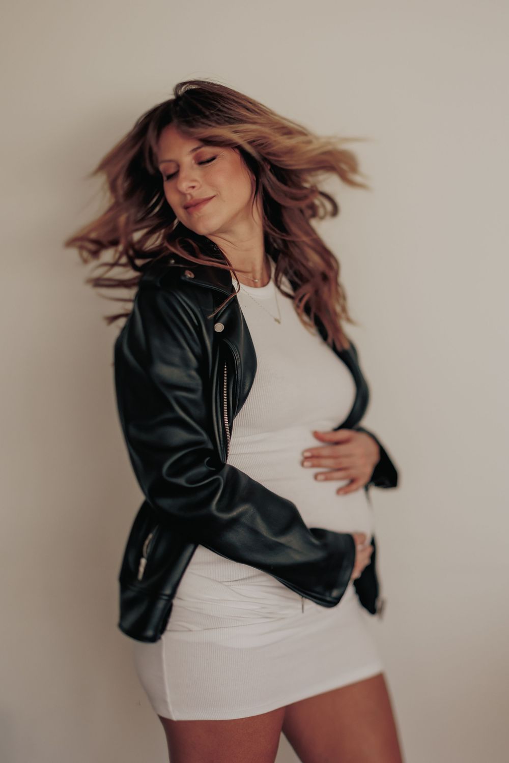 A person in a black leather jacket and white dress poses dynamically with windswept hair against a neutral wall.