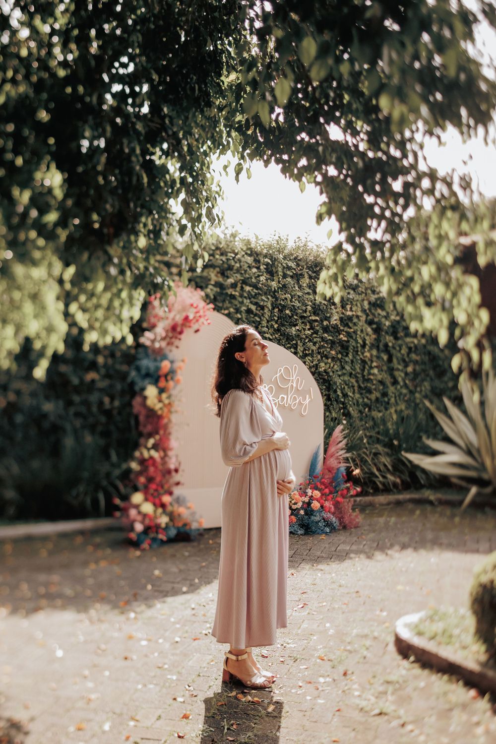 A figure in a flowing cream dress poses in front of a floral archway and greenery in a dreamy garden setting.