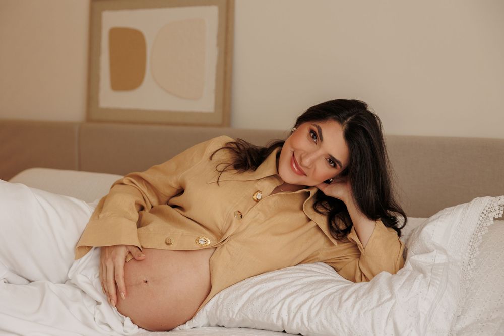 Person in beige sweater relaxing on white bedding in soft natural lighting.