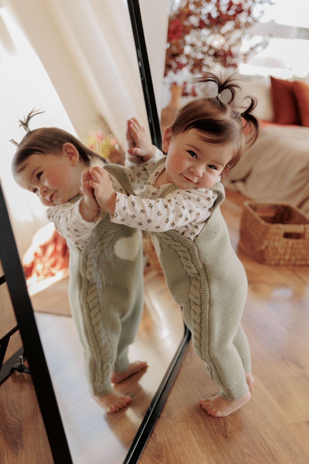 Baby in mint green knit romper looking at reflection in full-length mirror with playful expression.