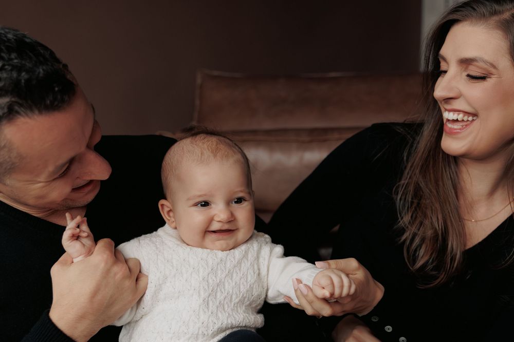 A baby in a white sweater sits between their parents on a brown couch while they all share a joyful moment together.