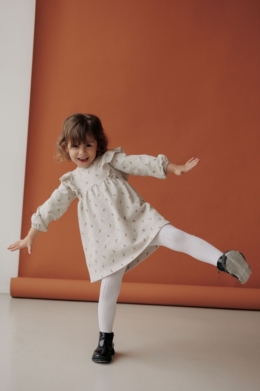Dancing child in white dress and tights strikes a playful pose against orange and white background.