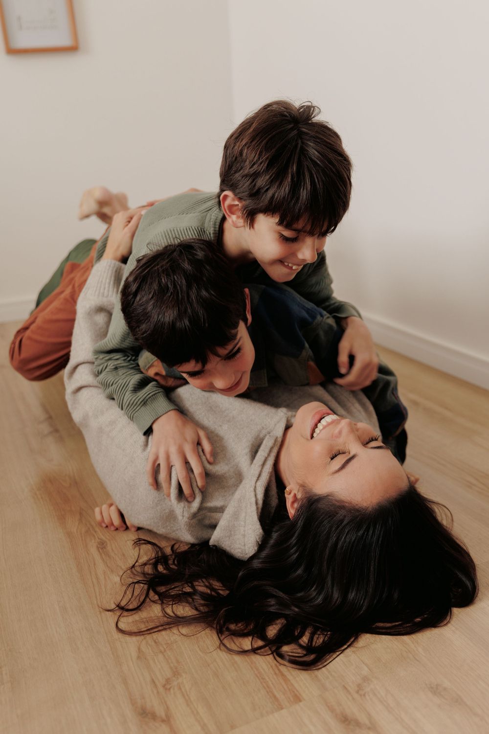 Three people lay playfully on a hardwood floor in a light-filled room sharing a joyful moment.