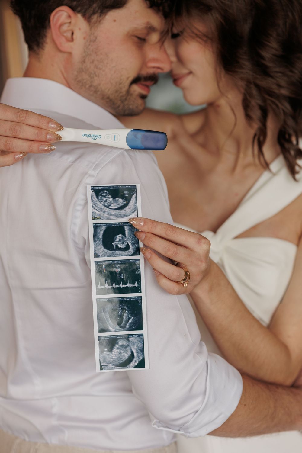 A couple in white clothing shares intimate moments while holding ultrasound photos of their expected baby.