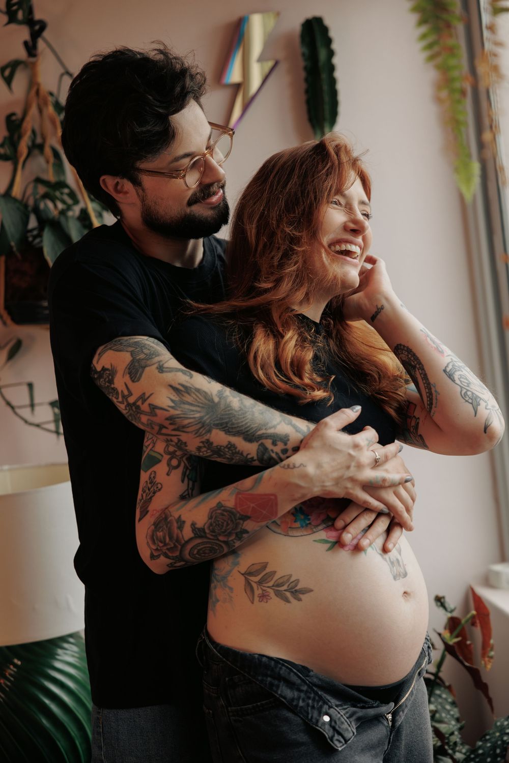 A tattooed pregnant person stands with their partner by a window, both smiling warmly among houseplants.