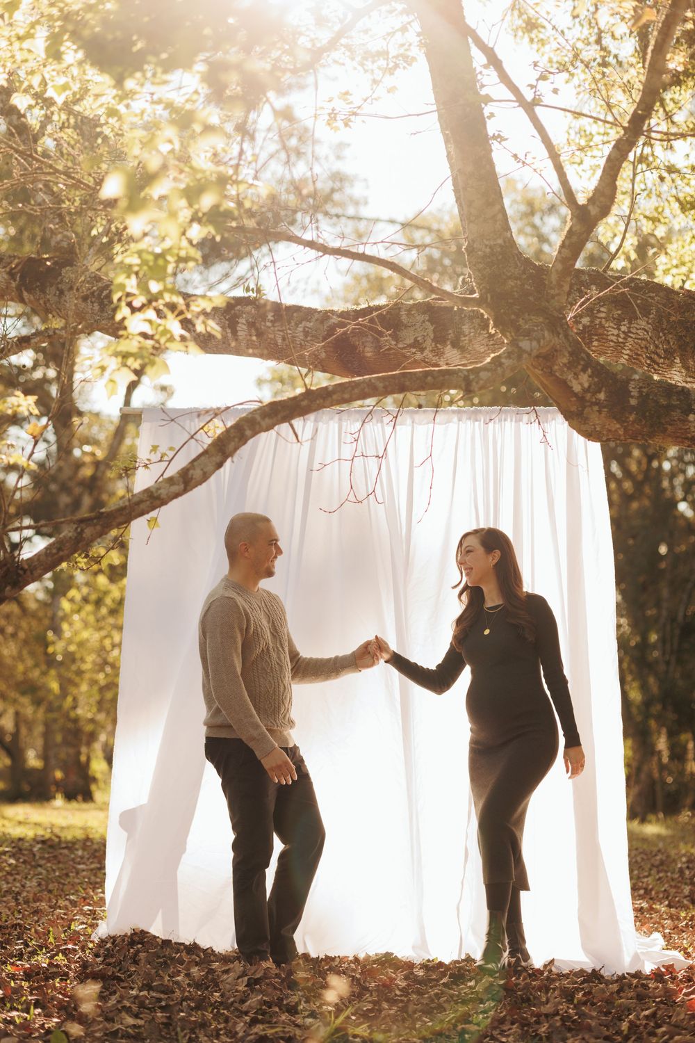 A romantic couple stands in front of white curtains hanging from tree branches in a sunlit autumn forest setting.