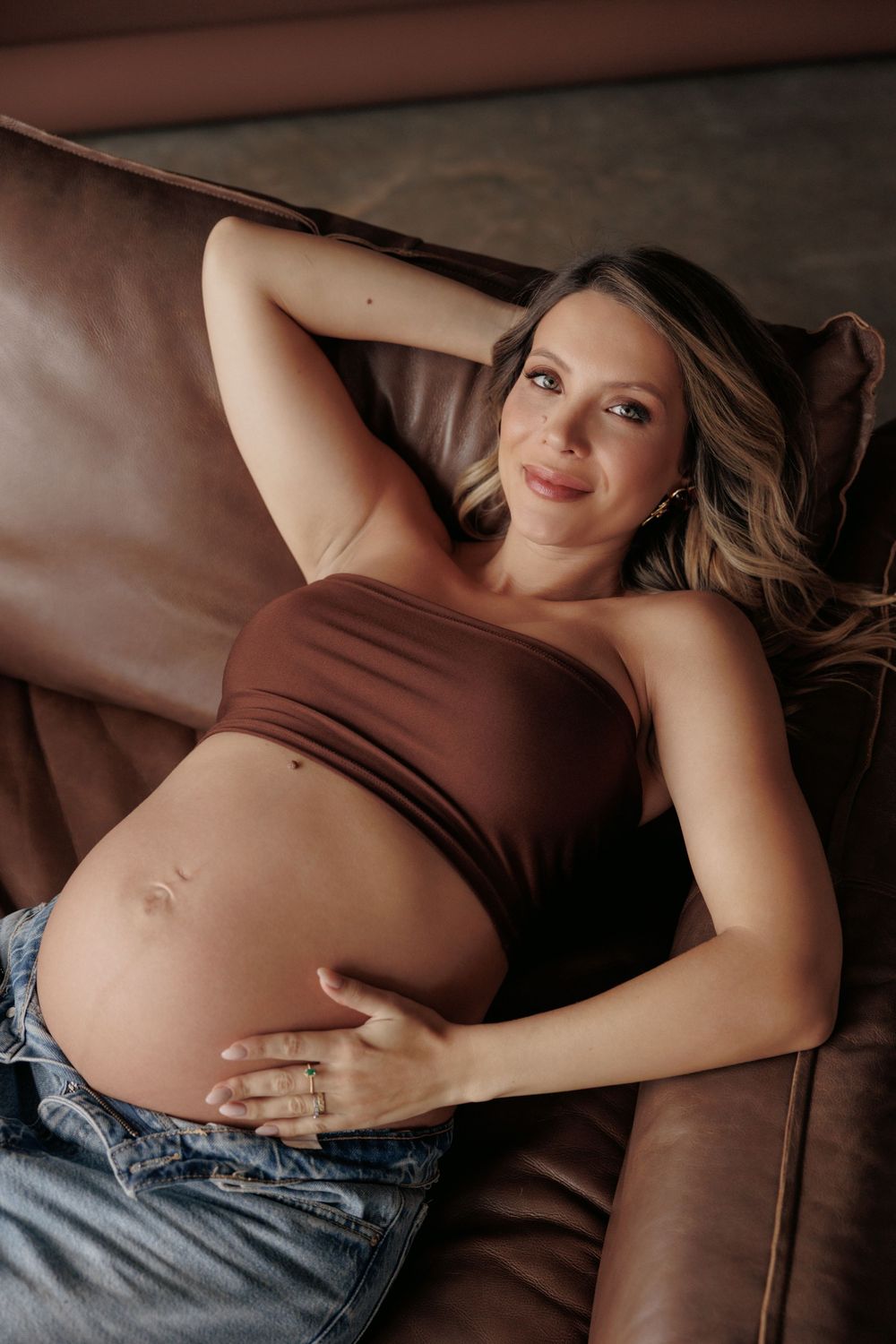 Pregnant woman in brown top and jeans reclines on brown leather couch with hands resting on her baby bump.