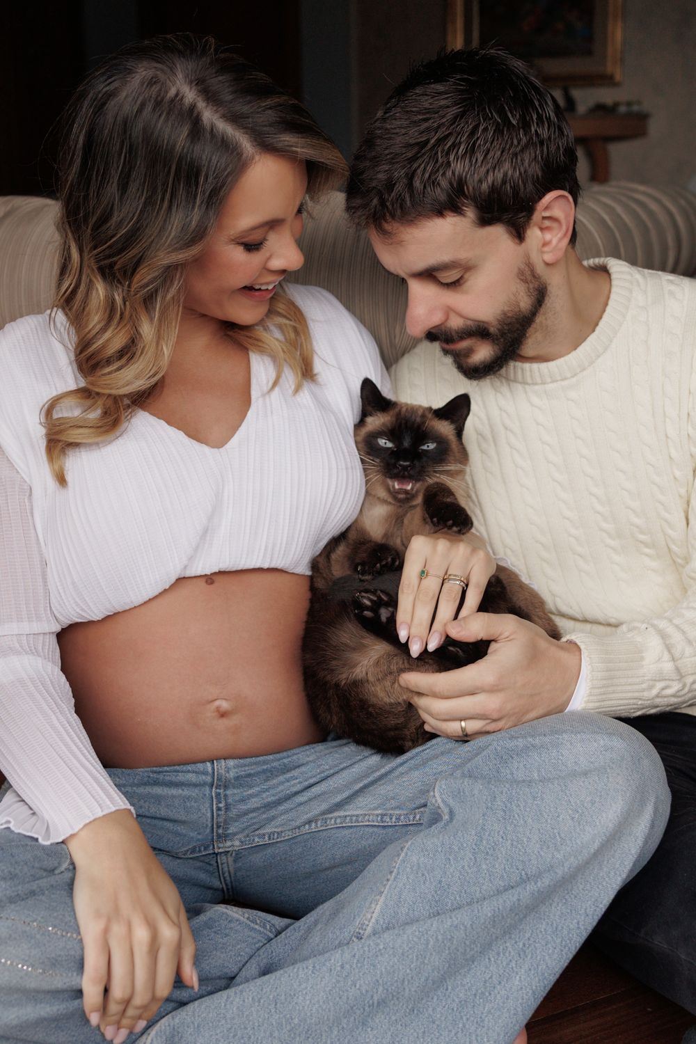 Happy couple in white sweaters sitting and playing with their Siamese cat on a couch at home.