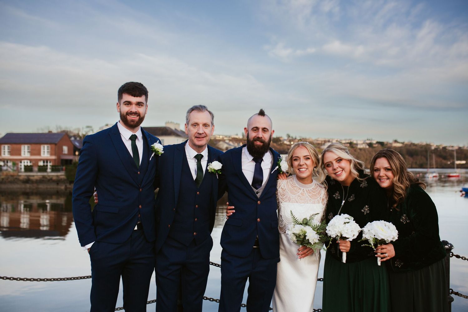 Wedding party poses together on a dock near waterfront houses under cloudy sky with bouquets.