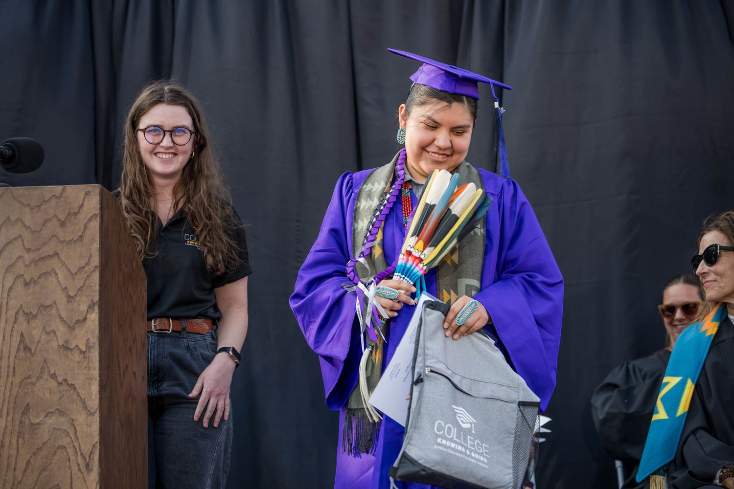 Graduate in bright purple cap and gown celebrates with diploma and backpack during outdoor ceremony.