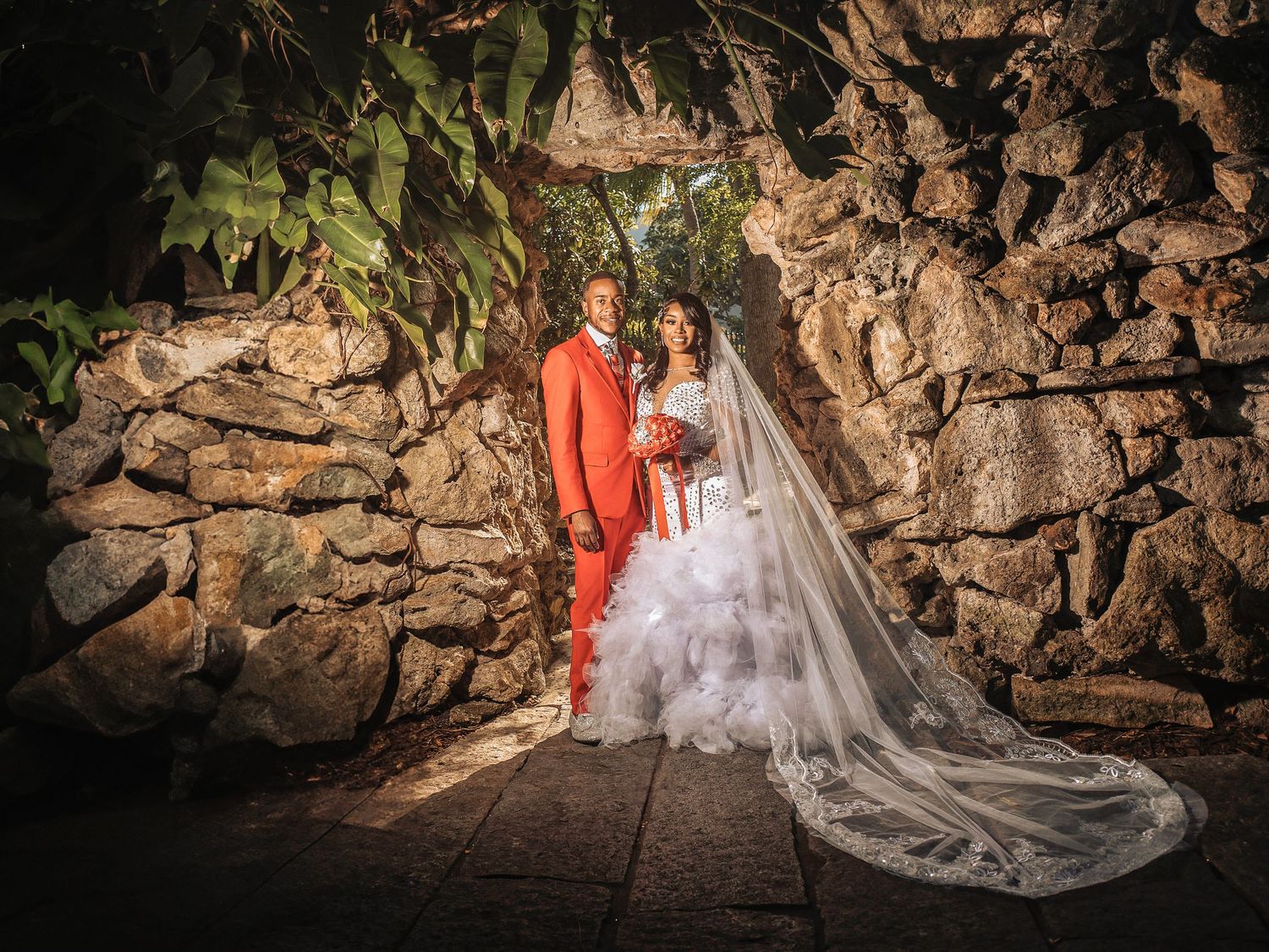 A couple poses in formal wedding attire against a rustic stone wall with dramatic lighting and trailing veil.