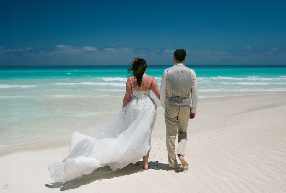 A couple in wedding attire walks along a pristine turquoise beach with white sand.