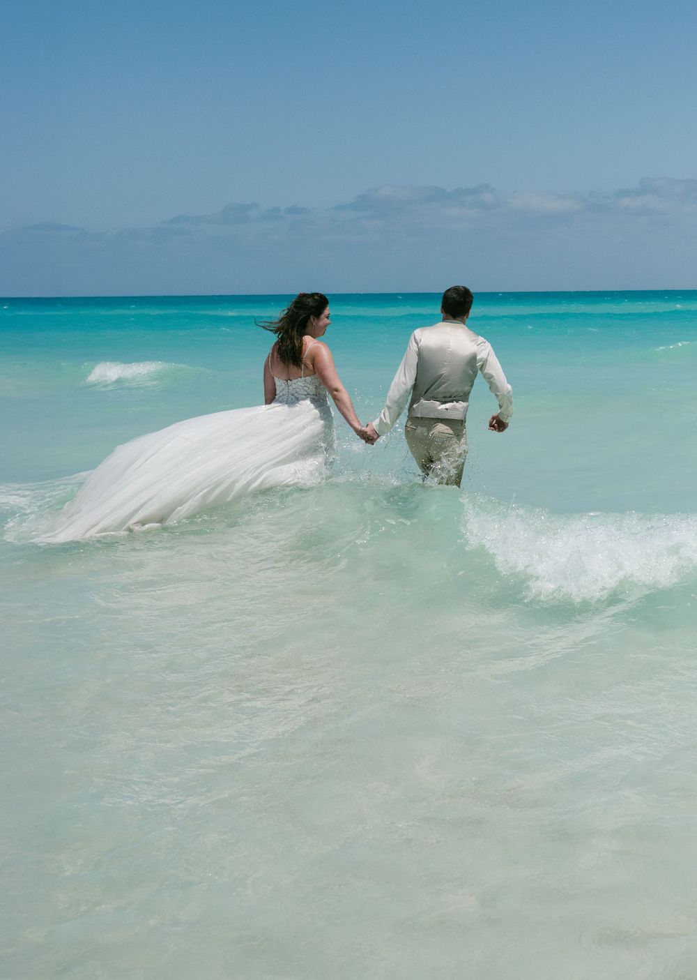 A couple in wedding attire wades through crystal clear turquoise ocean waters.