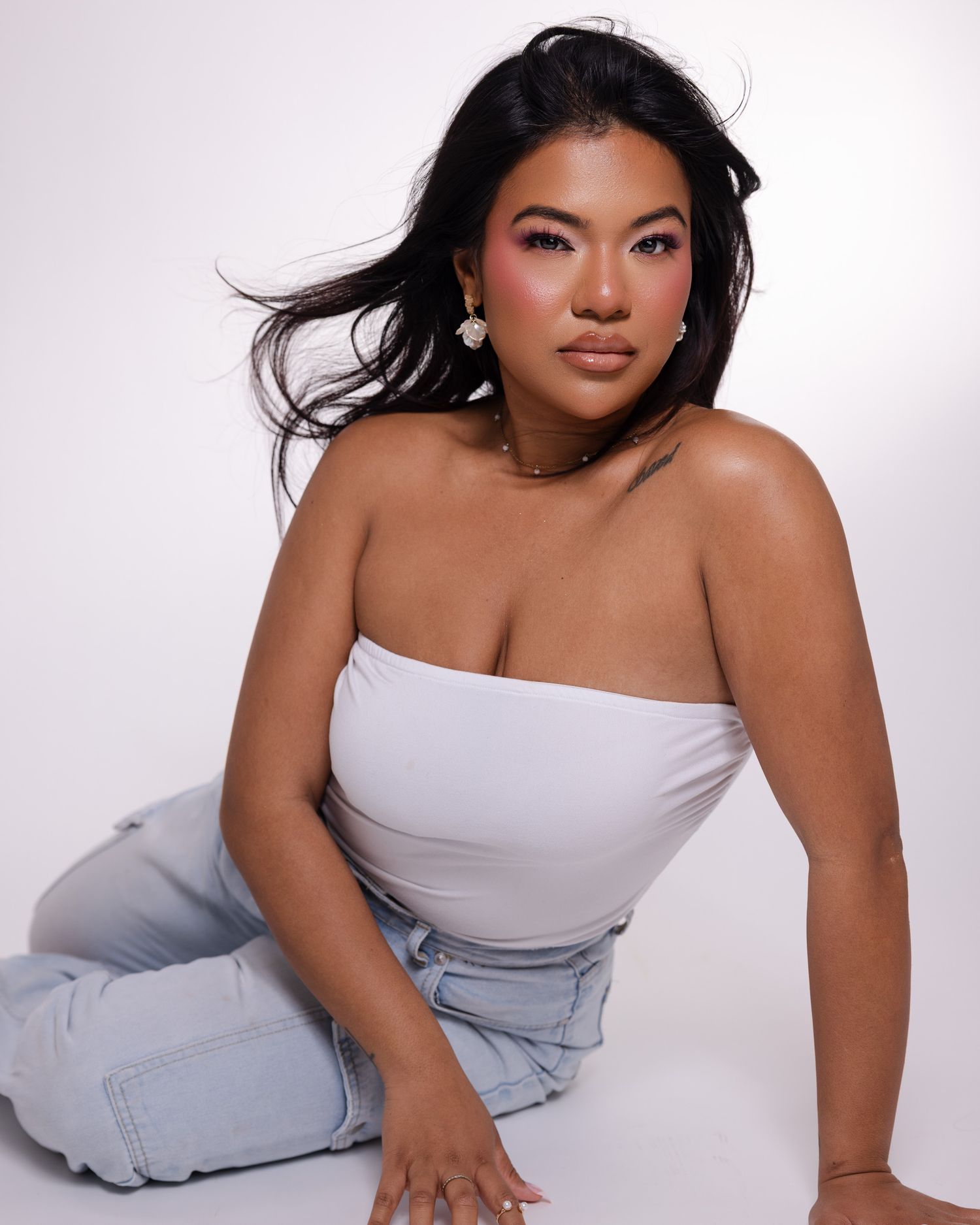 Glamour portrait of a woman in a white tube top and light jeans posing against a pale backdrop in a studio setting.