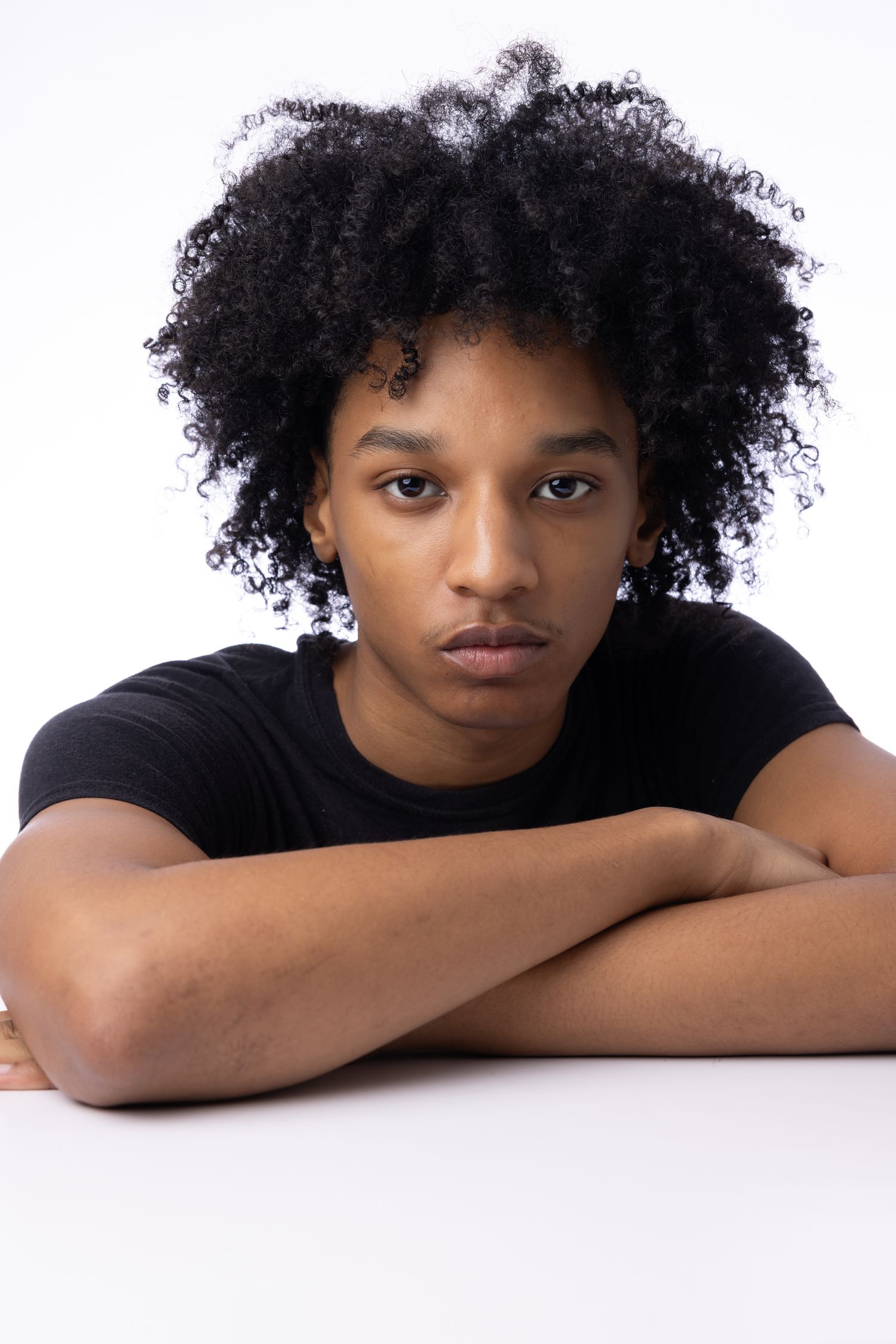 Young model with natural curly afro hairstyle wearing black t-shirt poses with arms crossed on white surface.