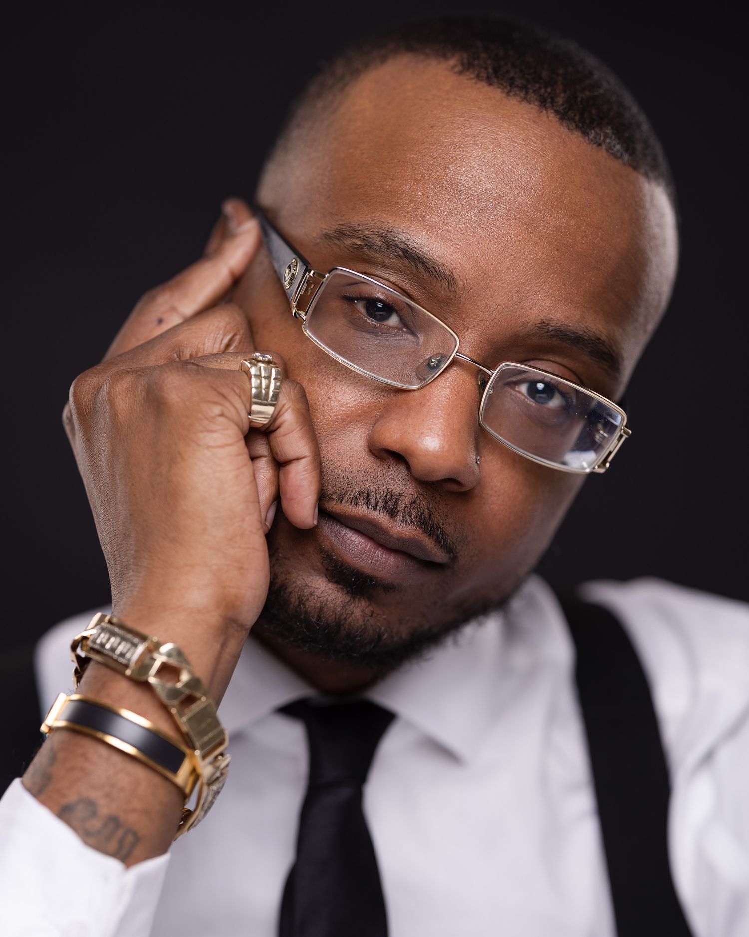 Professional portrait of a model in a white dress shirt, black tie and suspenders wearing glasses and gold jewelry.