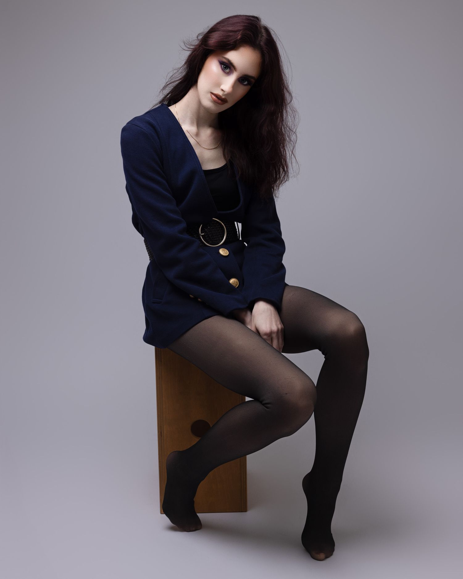A model in a navy blazer and black tights poses elegantly on a wooden stool against a grey studio backdrop.