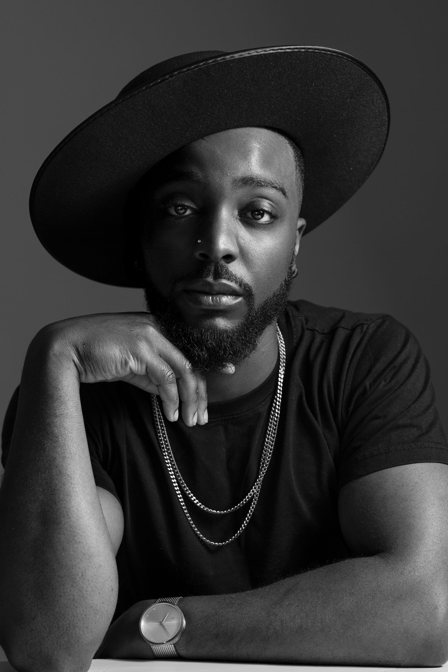 Black and white studio portrait of a model wearing a wide-brimmed hat, black t-shirt and silver chain necklace.