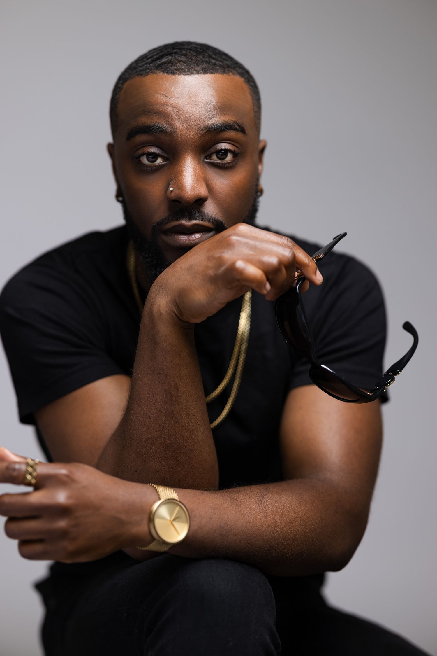 A male model wearing a black t-shirt and gold watch poses in front of a grey background with a thoughtful expression.
