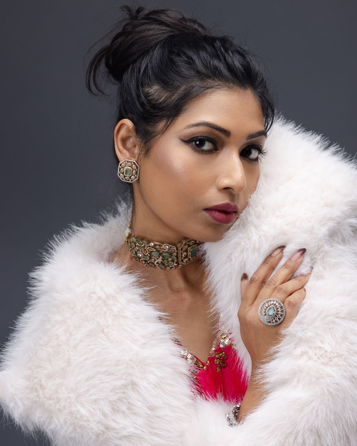 Model in glamorous white fur coat wearing ornate gold jewelry and red lips poses against dark backdrop.