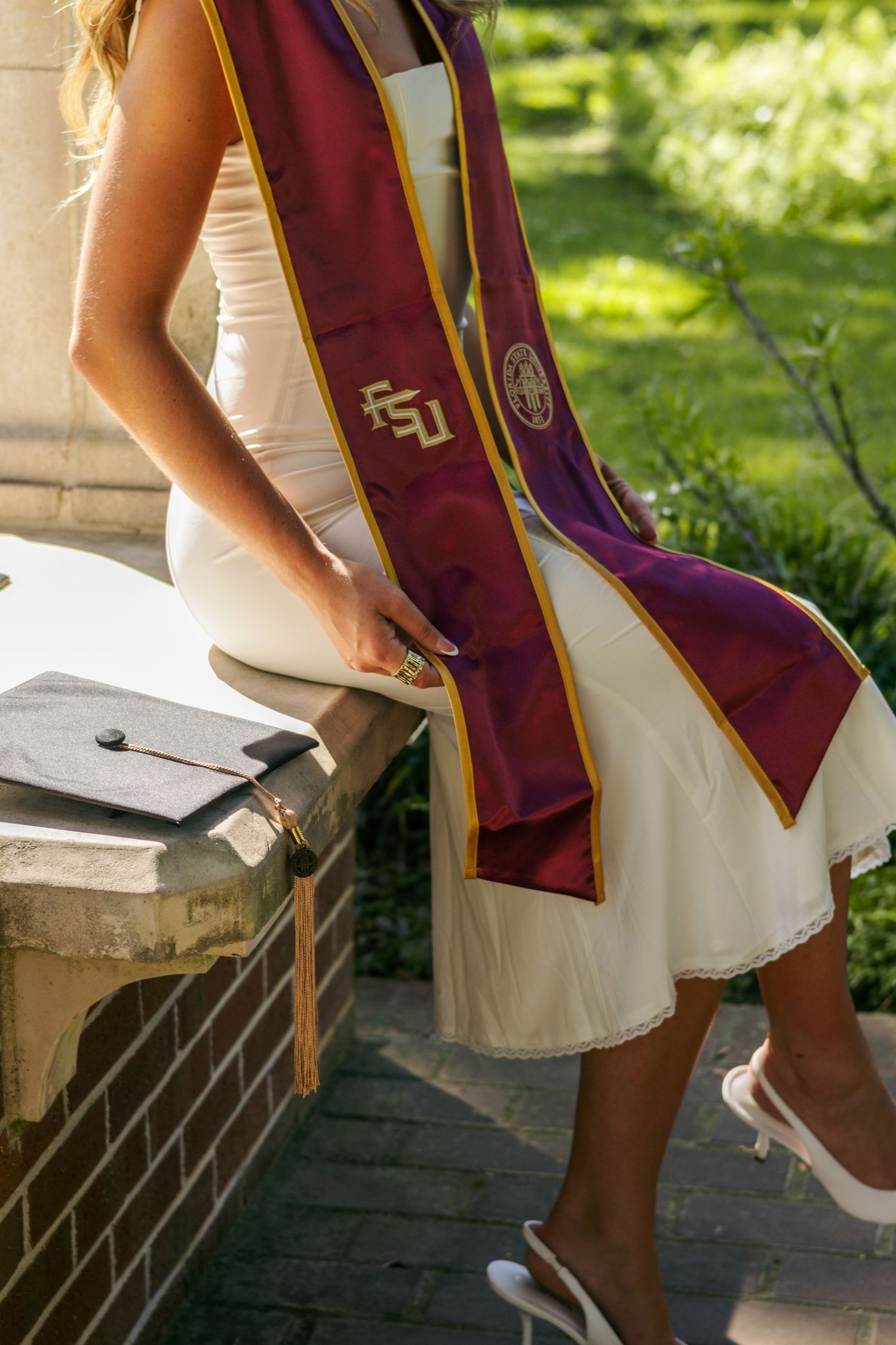 A detail photo of a girl with a garnet Florida State stole on, sitting on a brick and pavement wall, with her graduation cap beside her.