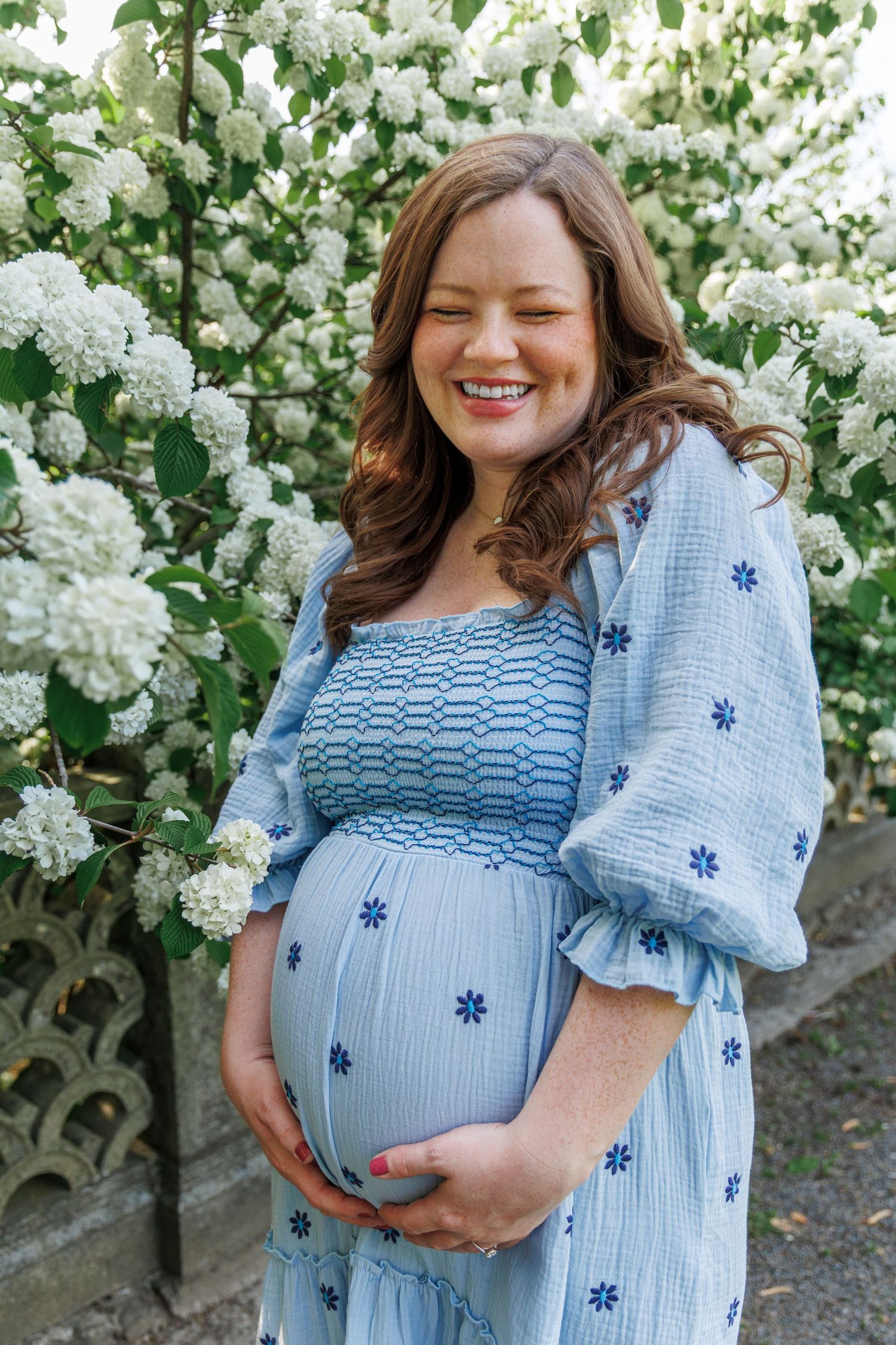 Maternity photos with blue smocked dress against blooming white flowers in garden setting.