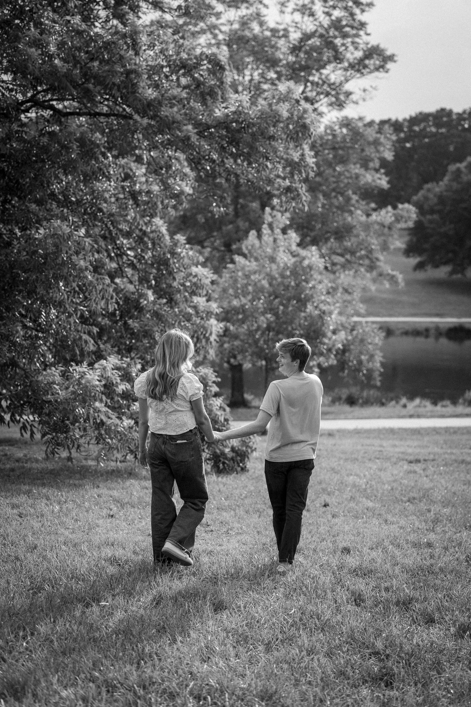 A black and white photo of two children holding hands while walking through a lush green park by a lake.