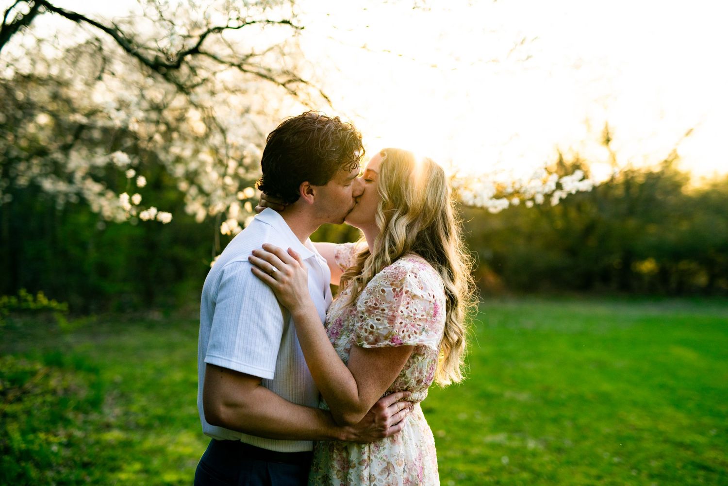 A romantic couple embraces and kisses in a sunlit meadow at golden hour with trees in the background.