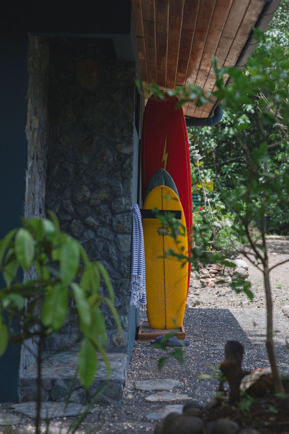 Vintage yellow and red surfboard leaning against stone wall with tropical foliage.
