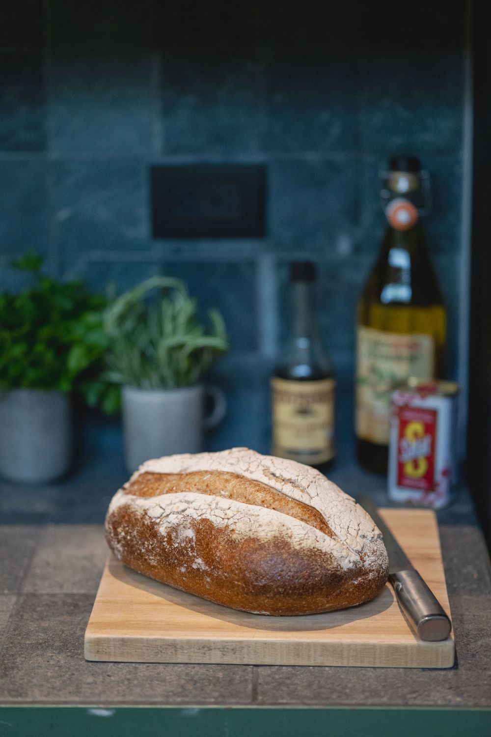 Artisanal sourdough bread loaf on wooden cutting board with herbs and bottles in background.