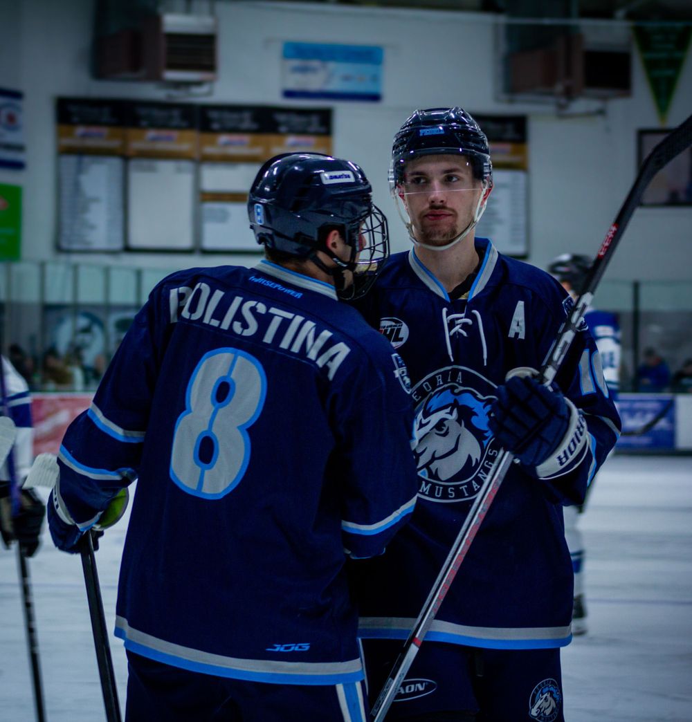 Two Peoria Mustangs Players Massimo Polistina and Zach Lacasse in blue jerseys discuss strategy during break in hockey game.